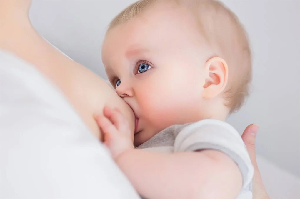 Ein blondes Baby mit blauen Augen trinkt an der mütterlichen Brust.