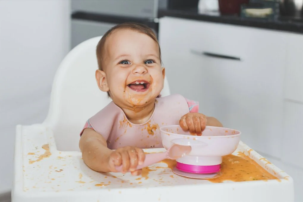 Ein fröhliches Baby sitzt gerade im Kinderstühlchen und ist Suppe aus einer Schüssel. Dabei hat sie gekleckert, hat eine schmutzige Schnute, hat einen Teil der Suppe verschüttet und freut sich sehr darüber.